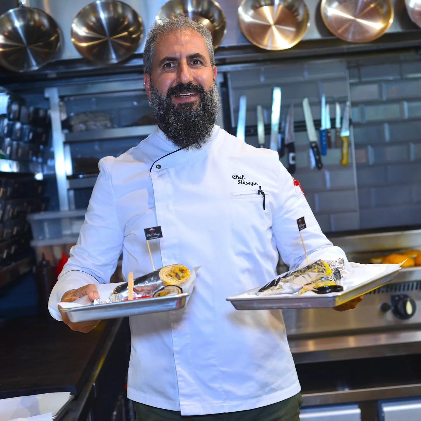 O Chef Hüseyin de jaleca branca na cozinha, com dois pratos na mão